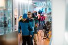 Ein Schüler und eine Lehrerin in blauer und schwarzer Kleidung gehen in einem hellen Flur mit Glasvitrinen, im Hintergrund weitere Menschen und ein Rollstuhl.